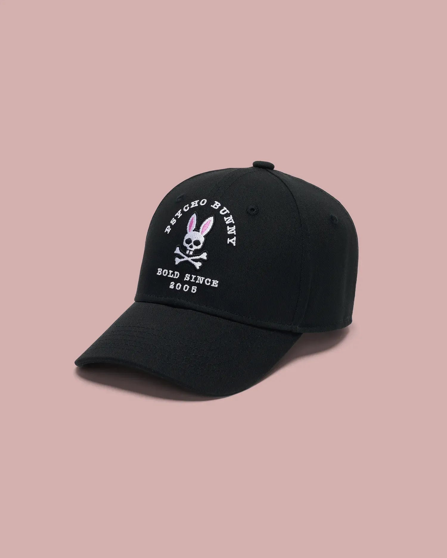 The KIDS ARCHIVE BASEBALL CAP - KO1500004 by Psycho Bunny is a black cotton twill cap with an embroidered bunny logo, pink ears and crossbones, and "PSYCHO BUNNY BOLD SINCE 2005," shown on a light pink background.