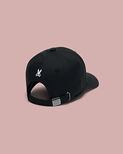The Psycho Bunny KIDS ARCHIVE BASEBALL CAP (KO1500004) is a black cotton twill cap shown from the back, featuring a small embroidered bunny logo above the adjustable strap with an engraved silver buckle. Background is solid light pink.