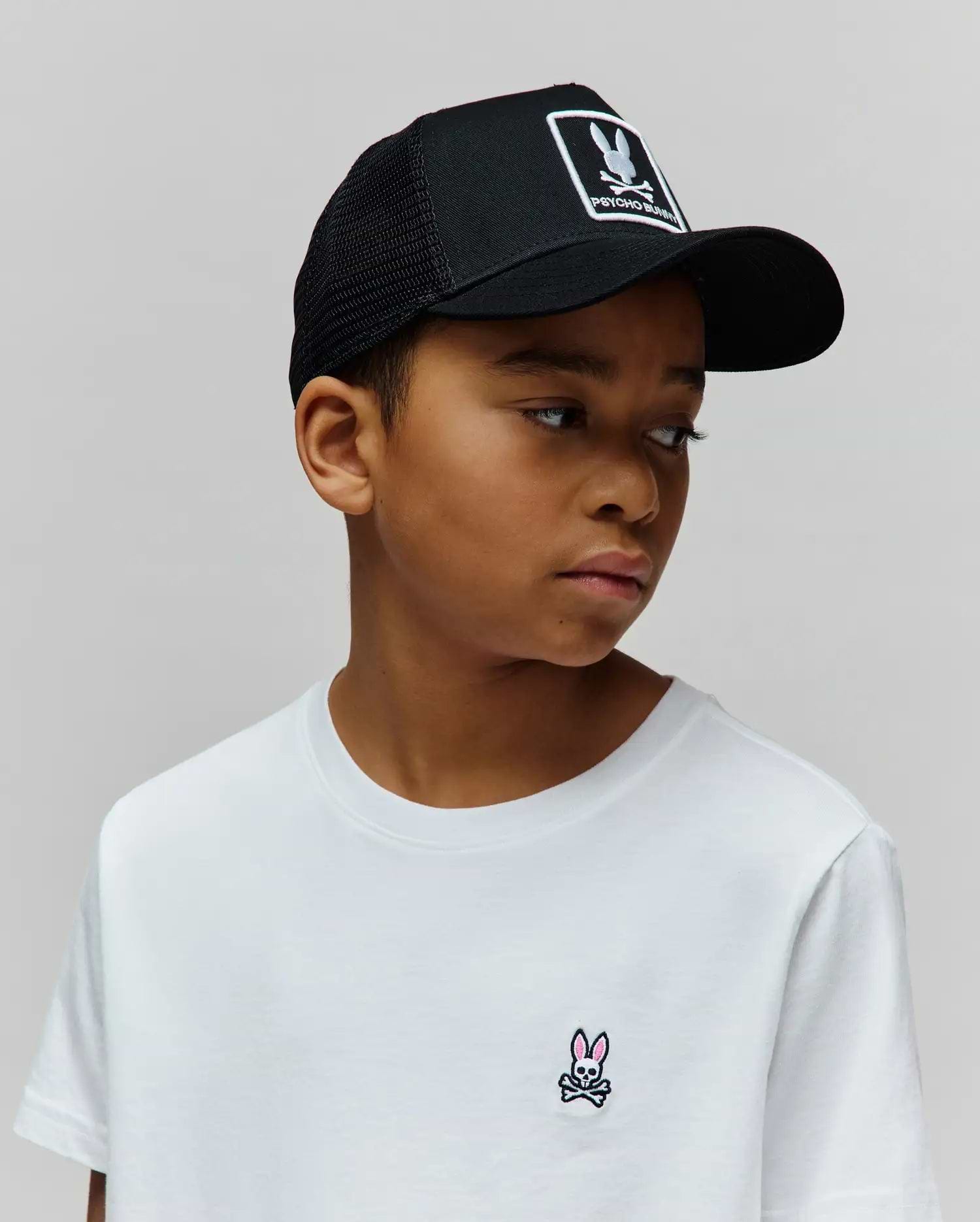 White jersey t-shirt with a small embroidered pink and black Psycho Bunny skull logo on the left chest, paired with a black mesh Kids Adamo Trucker Cap featuring a large white Psycho Bunny logo patch.