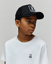White jersey t-shirt with a small embroidered pink and black Psycho Bunny skull logo on the left chest, paired with a black mesh Kids Adamo Trucker Cap featuring a large white Psycho Bunny logo patch.