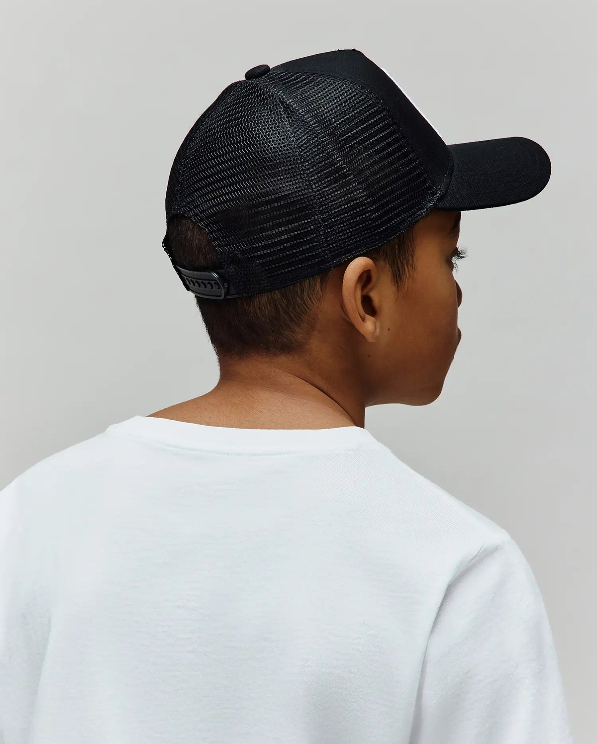 Black Kids Adamo Trucker Cap featuring a breathable mesh back, solid front panel, curved brim, and adjustable snapback closure, worn with a classic white relaxed-fit jersey crew neck t-shirt.