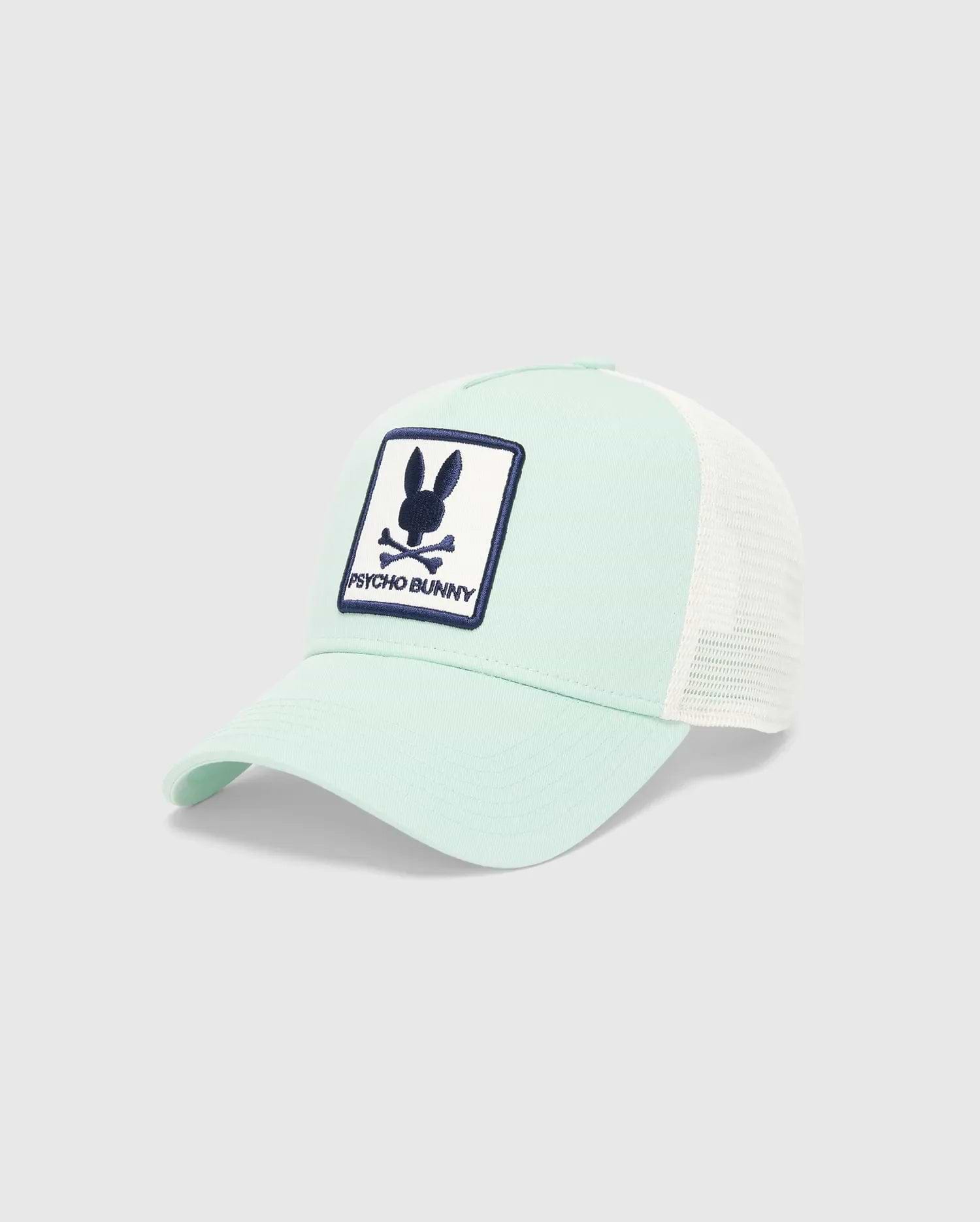 Kids Adamo Bunny Trucker Cap, a mint green cotton twill cap with a white mesh back. Features a prominent, embroidered navy blue and white Psycho Bunny skull logo patch on the front crown.