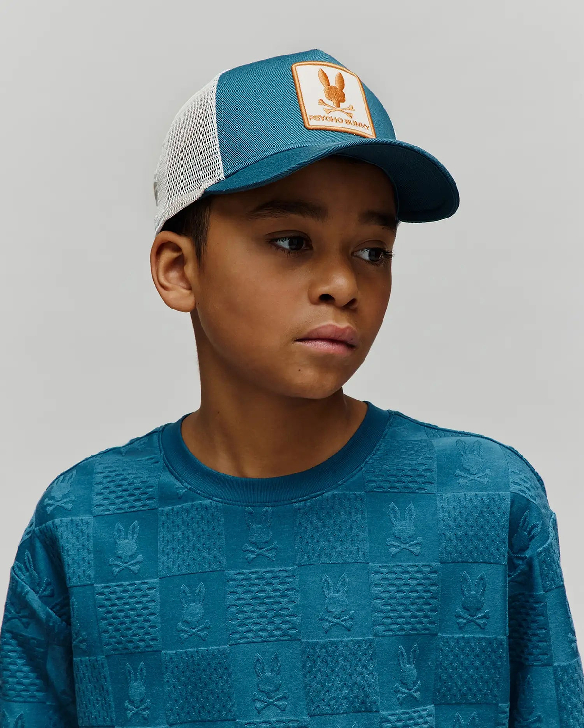 KIDS ADAMO TRUCKER CAP in teal with white mesh back and an orange Psycho Bunny logo patch, paired with a teal crewneck sweatshirt featuring an all-over embossed checkerboard pattern of Psycho Bunny skull logos.