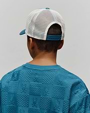 Kids Adamo Trucker Cap with a teal brim and white mesh back, paired with a teal blue relaxed-fit sweatshirt featuring an all-over embossed checkerboard pattern of Psycho Bunny skull logos and textured squares.