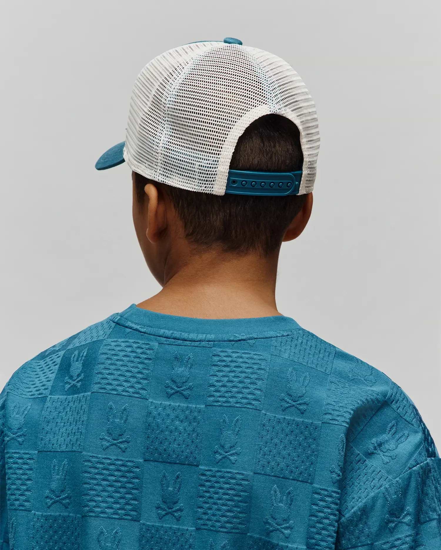 Kids Adamo Trucker Cap with a teal brim and white mesh back, paired with a teal blue relaxed-fit sweatshirt featuring an all-over embossed checkerboard pattern of Psycho Bunny skull logos and textured squares.