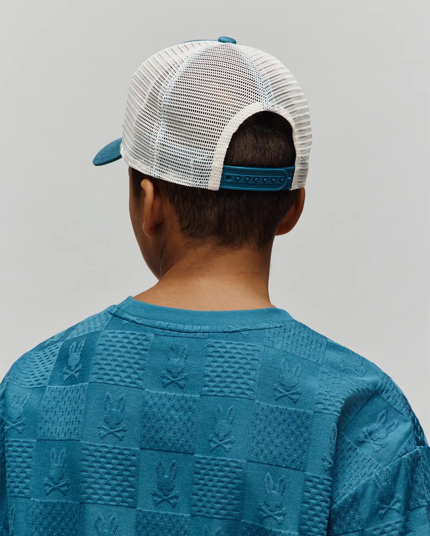 Kids Adamo Trucker Cap with a teal brim and white mesh back, paired with a teal blue relaxed-fit sweatshirt featuring an all-over embossed checkerboard pattern of Psycho Bunny skull logos and textured squares.