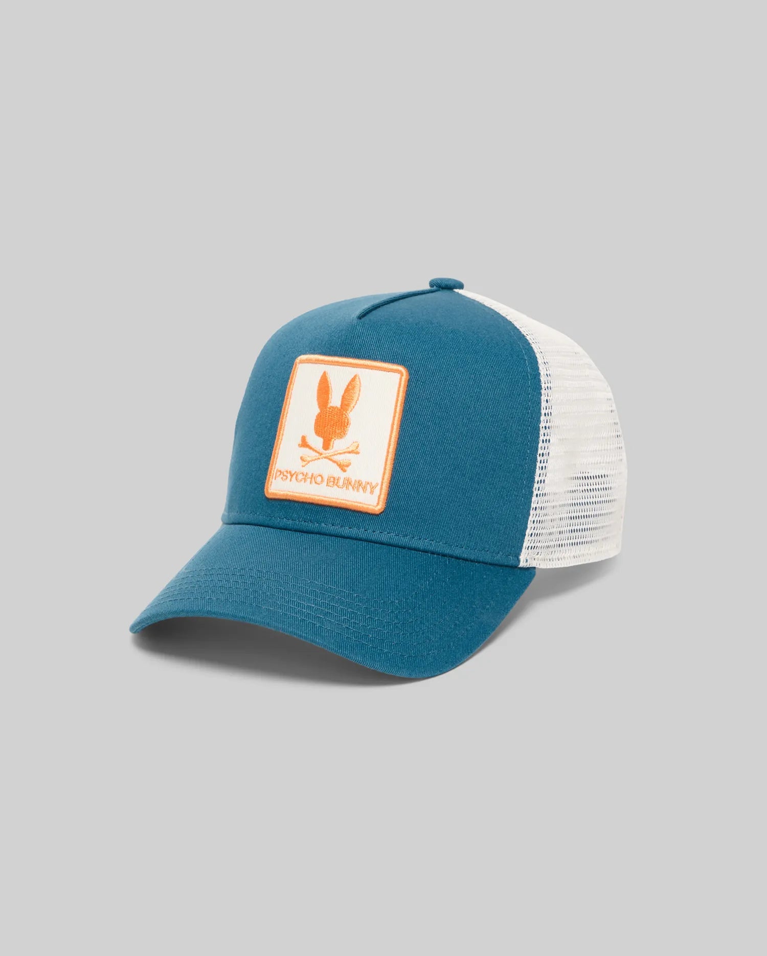 Kids Adamo Trucker Cap in teal blue with white mesh back. Features a large, embroidered orange Psycho Bunny skull logo patch on a cream background on the front crown.