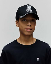 Black jersey crew neck t-shirt with a small pink and white embroidered Psycho Bunny logo on the left chest, paired with a black Kids Luke Trucker Cap featuring a large white embroidered Psycho Bunny skull logo and patterned brim.