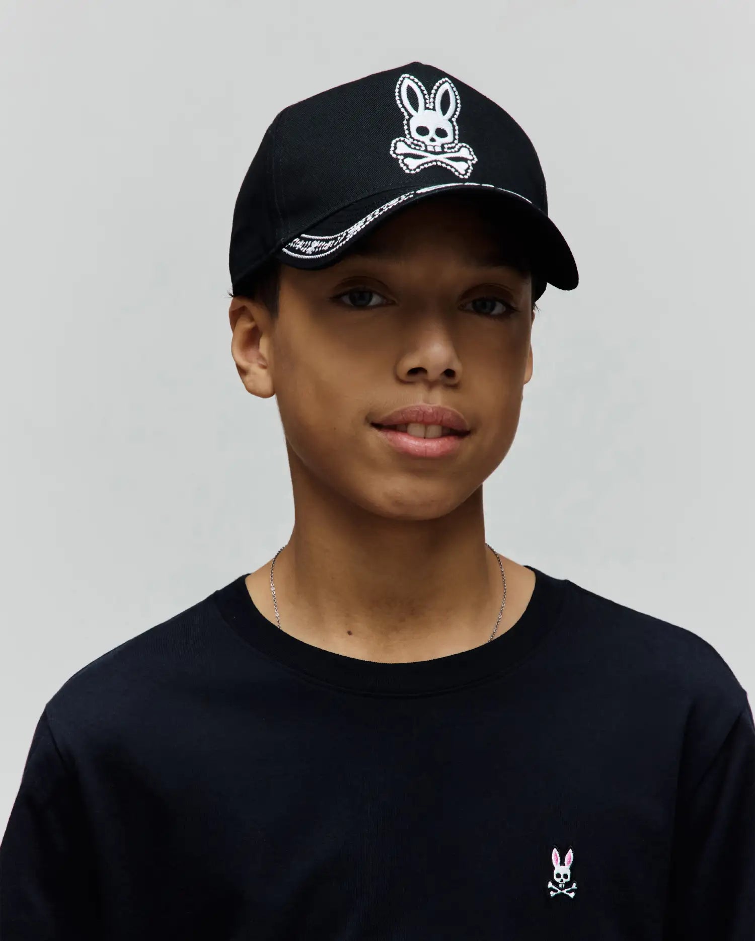 Black jersey crew neck t-shirt with a small pink and white embroidered Psycho Bunny logo on the left chest, paired with a black Kids Luke Trucker Cap featuring a large white embroidered Psycho Bunny skull logo and patterned brim.