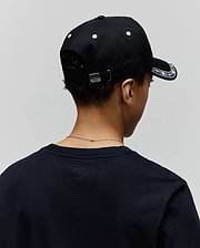 Black Kids Luke Trucker Cap with white embroidered brim pattern and eyelets, featuring a silver Psycho Bunny skull logo on the back buckle, paired with a dark navy classic fit jersey t-shirt.
