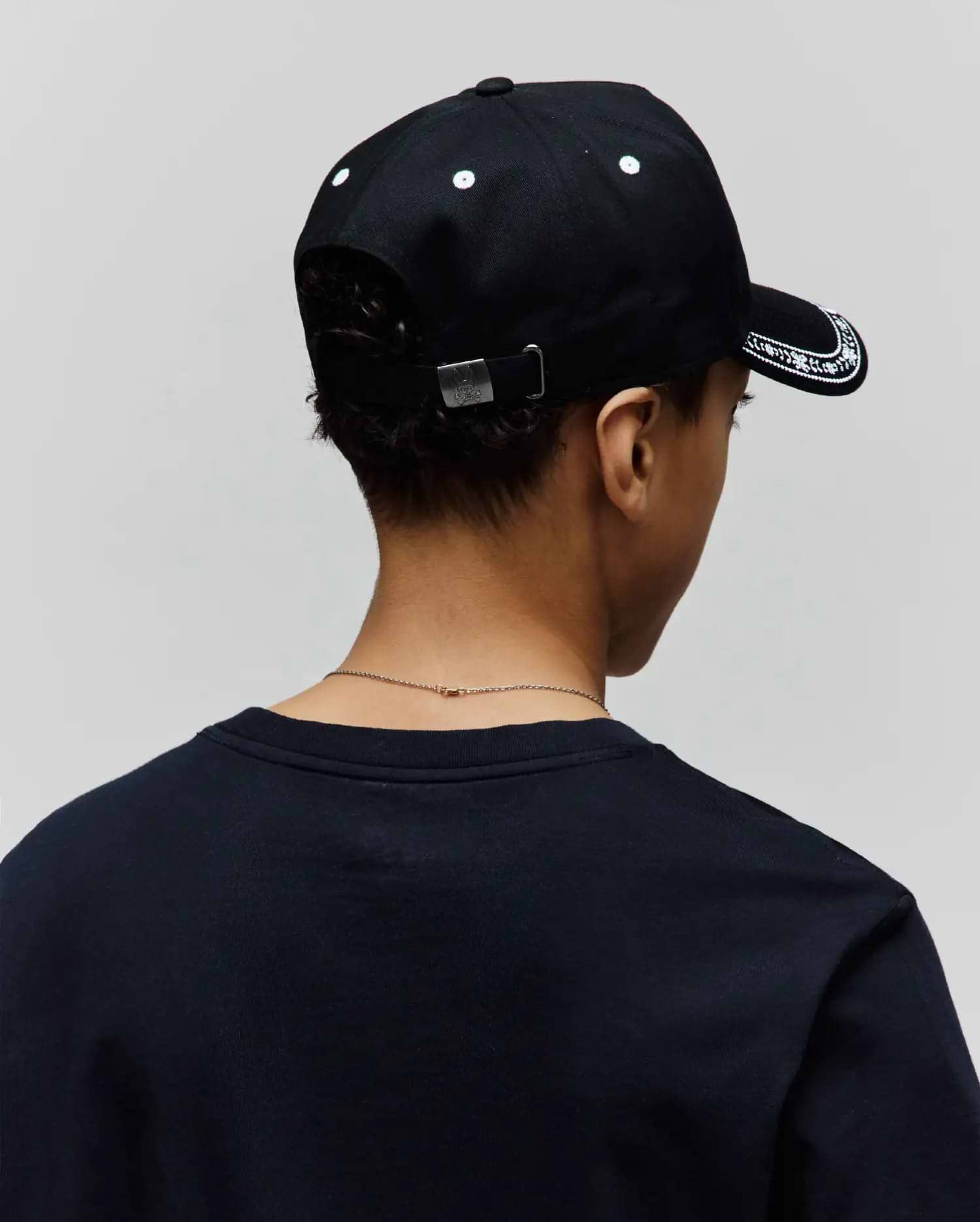 Black Kids Luke Trucker Cap with white embroidered brim pattern and eyelets, featuring a silver Psycho Bunny skull logo on the back buckle, paired with a dark navy classic fit jersey t-shirt.