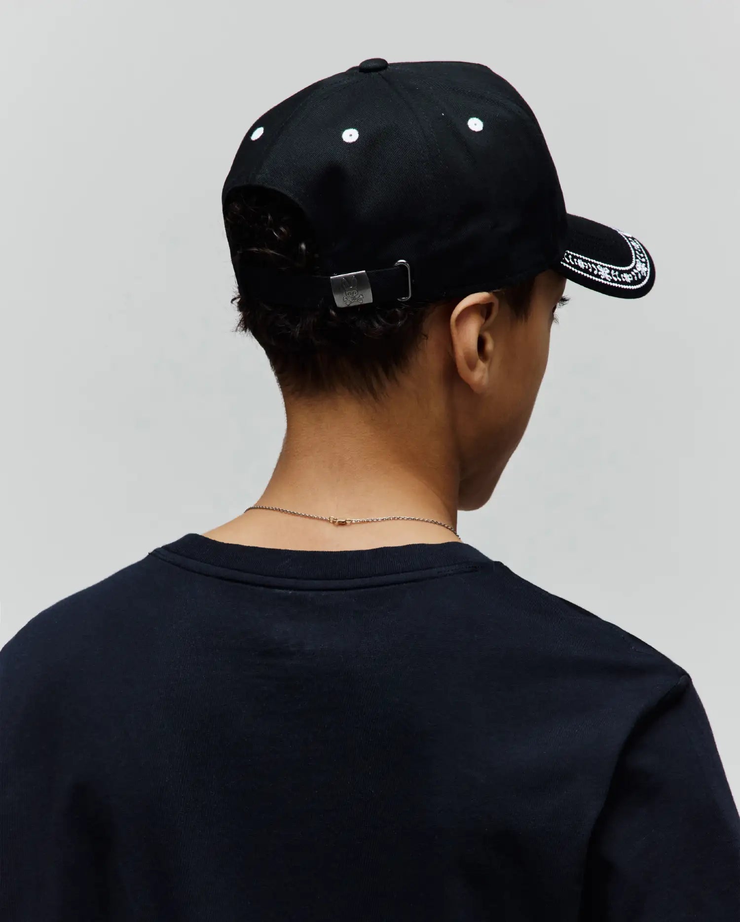 Black Kids Luke Trucker Cap with white embroidered brim pattern and eyelets, featuring a silver Psycho Bunny skull logo on the back buckle, paired with a dark navy classic fit jersey t-shirt.