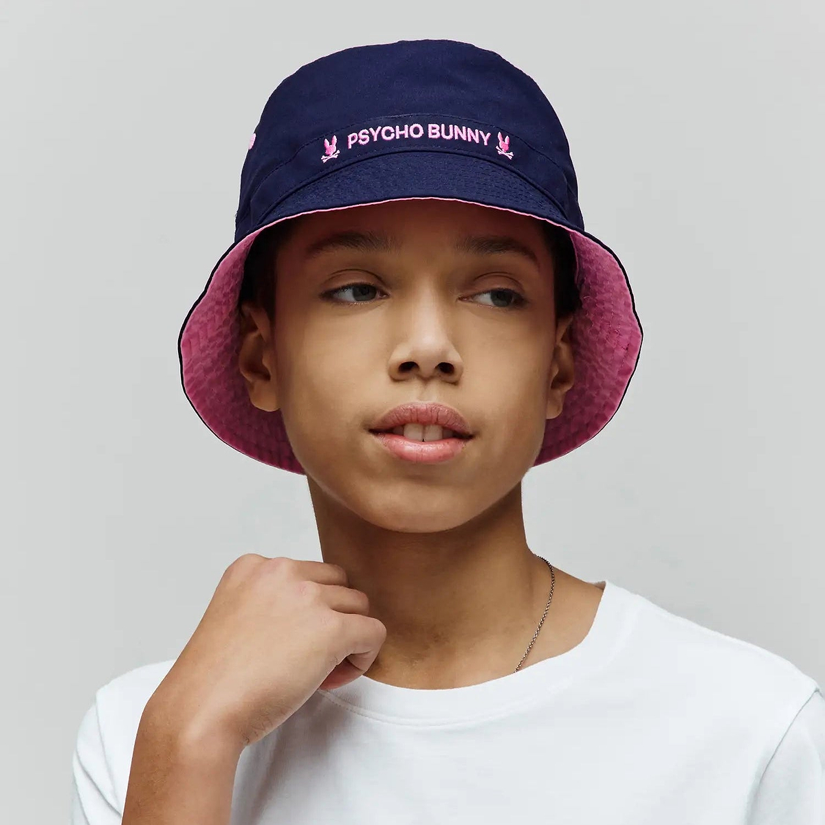 Kids Luke Reversible Bucket Hat in Navy Blue, reversing to Pink, with pink embroidered Psycho Bunny text and skull logos, plus a white jersey t-shirt featuring a small embroidered Psycho Bunny skull logo.