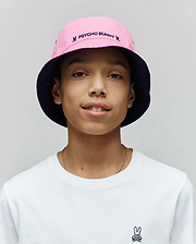 White classic fit jersey t-shirt with a small embroidered Psycho Bunny skull logo on the left chest, paired with a Kids Luke Reversible Bucket Hat in pink with a navy underside, featuring embroidered Psycho Bunny text and small bunny skull logos.