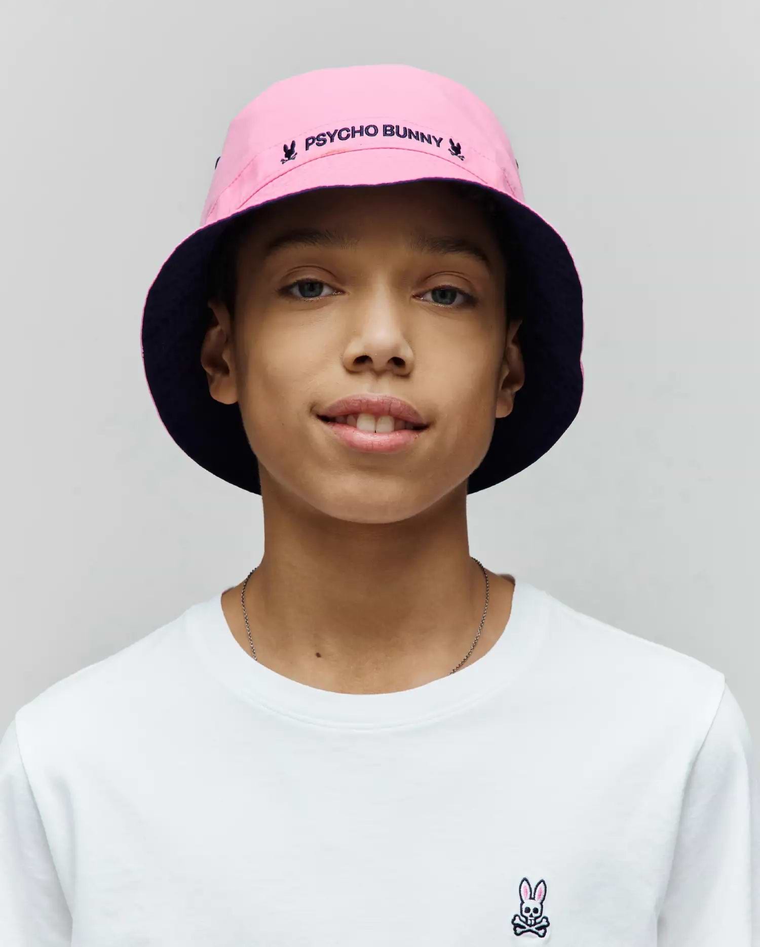 White classic fit jersey t-shirt with a small embroidered Psycho Bunny skull logo on the left chest, paired with a Kids Luke Reversible Bucket Hat in pink with a navy underside, featuring embroidered Psycho Bunny text and small bunny skull logos.