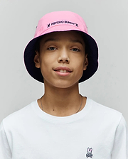 White classic fit jersey t-shirt with a small embroidered Psycho Bunny skull logo on the left chest, paired with a Kids Luke Reversible Bucket Hat in pink with a navy underside, featuring embroidered Psycho Bunny text and small bunny skull logos.