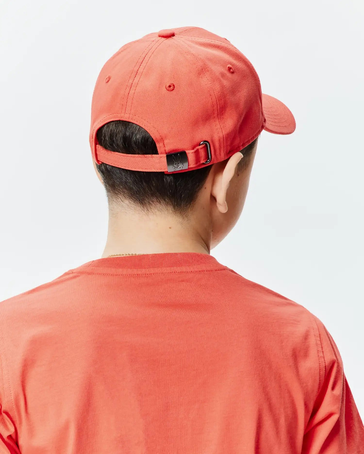 Vibrant coral KIDS CRAIG BASEBALL CAP, featuring a small black Psycho Bunny skull logo on its adjustable metal buckle, complementing a relaxed fit coral jersey t-shirt.