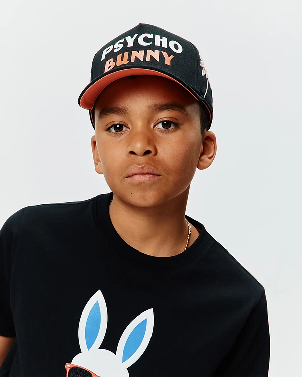 Black KIDS BRAD TRUCKER CAP with orange brim and white/orange PSYCHO BUNNY text, paired with a black jersey t-shirt featuring a large white bunny skull graphic with blue ears and orange sunglasses.