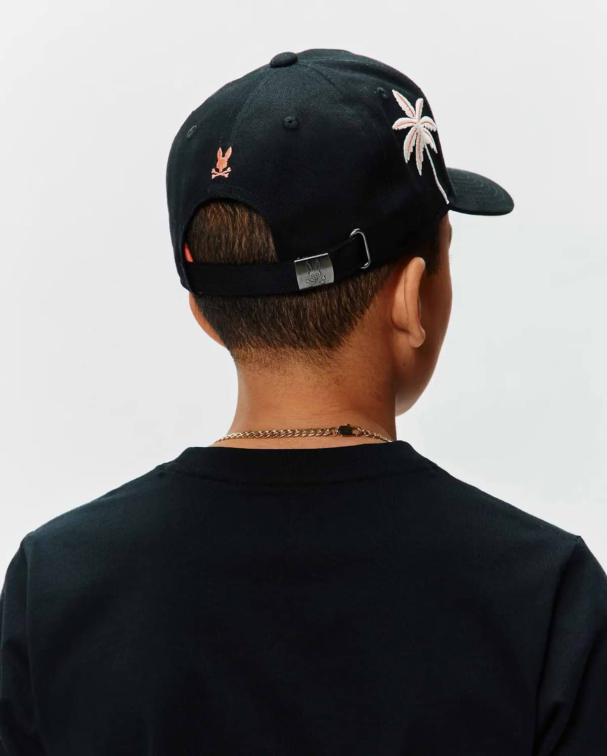 Black Kids Brad Trucker Cap featuring an embroidered orange Psycho Bunny skull logo on the back, a white and pink palm tree graphic on the side, and a metal buckle with an engraved bunny skull.
