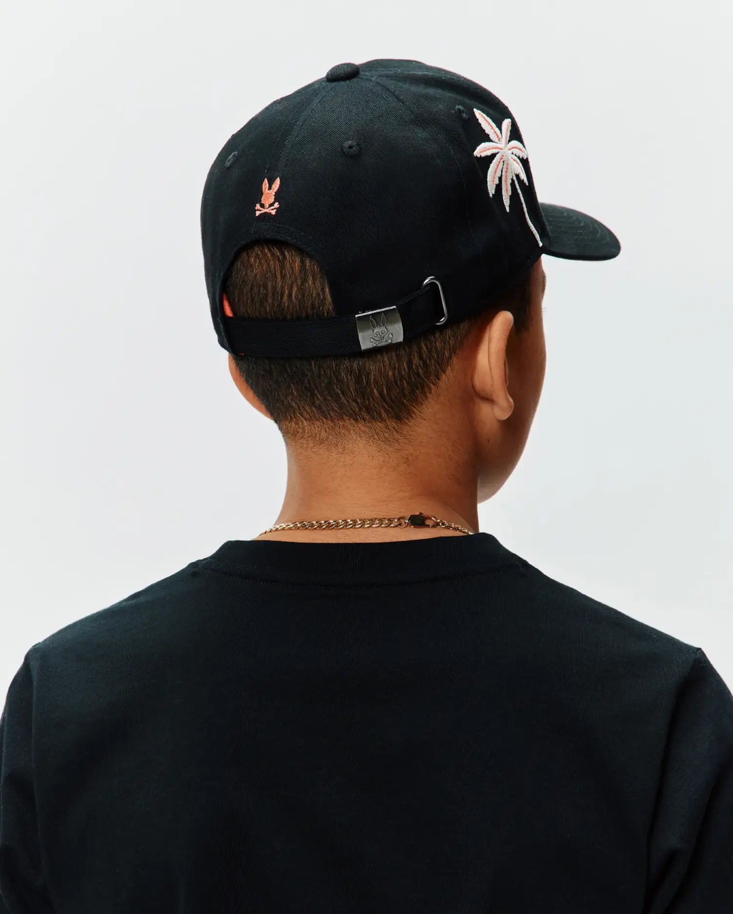 Black Kids Brad Trucker Cap featuring an embroidered orange Psycho Bunny skull logo on the back, a white and pink palm tree graphic on the side, and a metal buckle with an engraved bunny skull.
