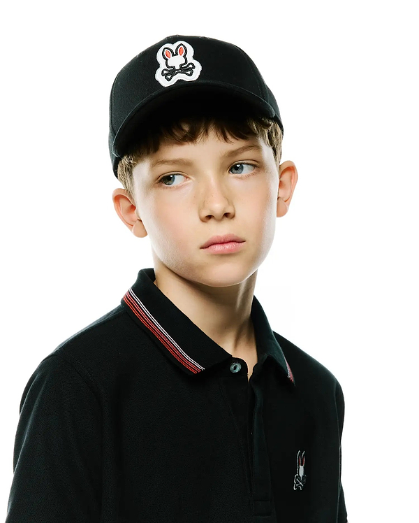 Black pique polo with red and white striped collar, small embroidered Psycho Bunny logo on chest, and a Black Kids Jaydon Baseball Cap with a large embroidered bunny skull logo.