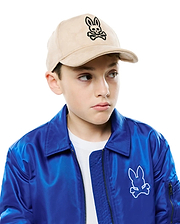 Beige suede Kids Kenneth A-Frame Trucker Cap with a black embroidered Psycho Bunny logo on the front, and a vibrant royal blue satin bomber jacket featuring a large white embroidered Psycho Bunny logo on the left chest.