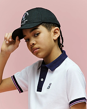 A young person with braided hair wears the Psycho Bunny KIDS ARCHIVE BASEBALL CAP - KO1500004 in black cotton twill and a white polo with navy trim, standing against a pink background while slightly lifting the cap's brim.