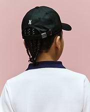 A child with braided hair wears the Psycho Bunny KIDS ARCHIVE BASEBALL CAP - KO1500004, a black cotton twill hat with an embroidered bunny logo, paired with a white shirt and dark blue collar, set against a light pink background.