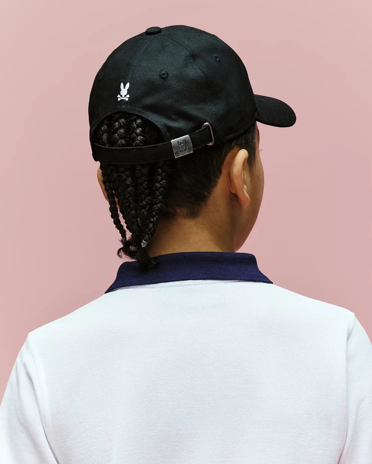 A child with braided hair wears the Psycho Bunny KIDS ARCHIVE BASEBALL CAP - KO1500004, a black cotton twill hat with an embroidered bunny logo, paired with a white shirt and dark blue collar, set against a light pink background.