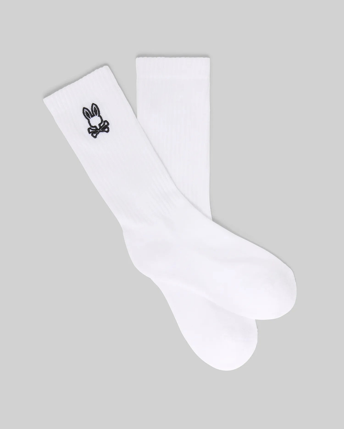 The Psycho Bunny KIDS 3 PACK SPORT SOCK (KO2200001) includes white crew socks made with soft Pima cotton and spandex, each featuring a small black bunny skull and crossbones logo near the top. Displayed on a gray background.
