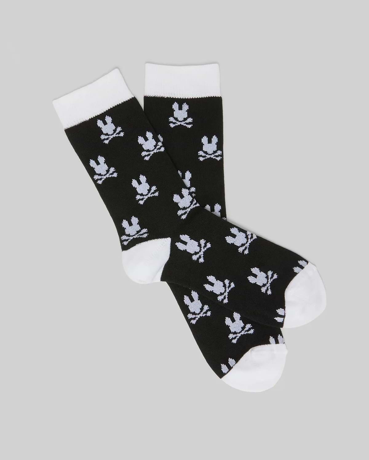 The Psycho Bunny KIDS 2 PACK ALL OVER BUNNY DRESS SOCK (KO2200003) features black socks with white toes, heels, and cuffs. A bunny skull-and-crossbones pattern is knit into soft stretch Pima cotton blend, shown on a light gray background.