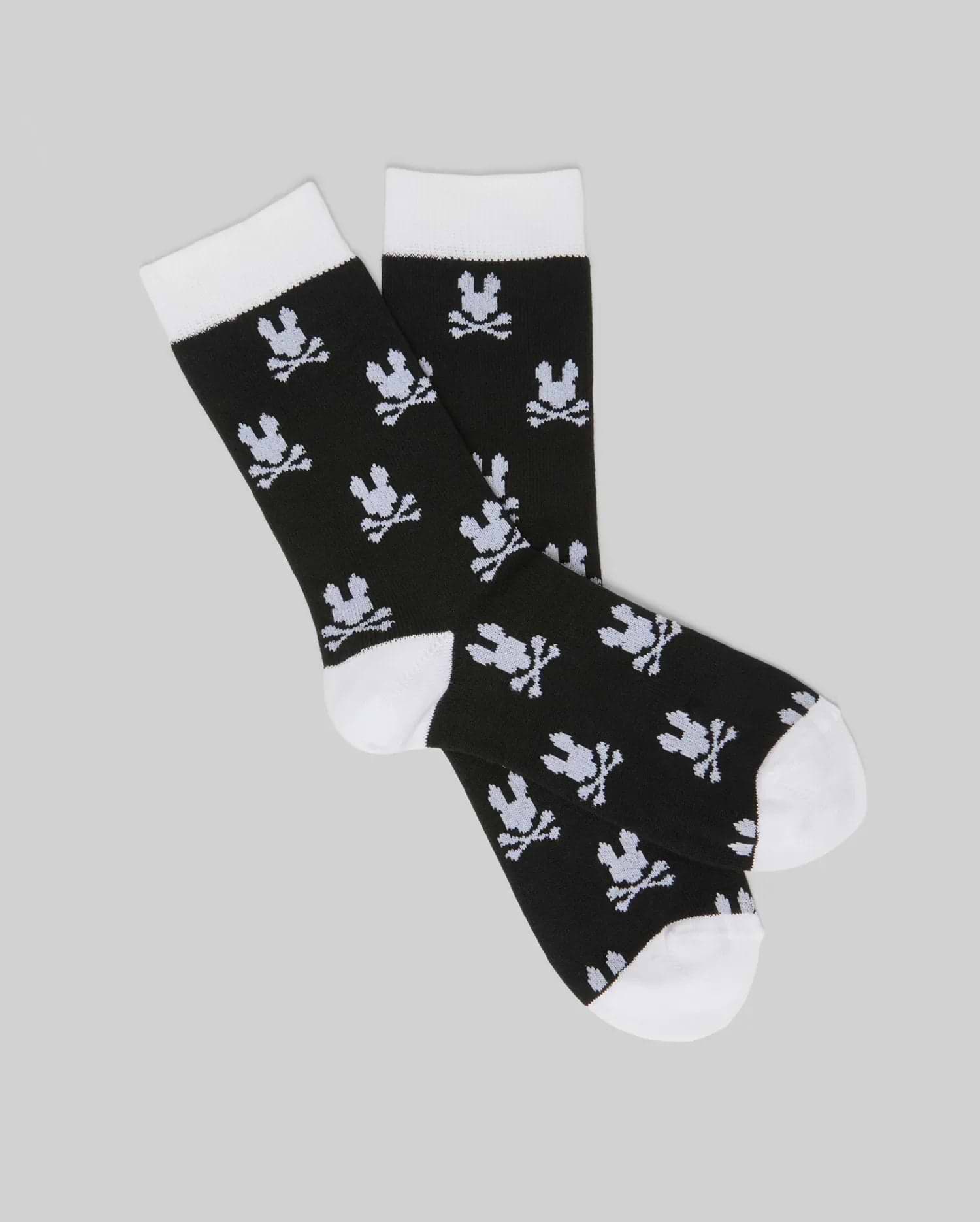 The Psycho Bunny KIDS 2 PACK ALL OVER BUNNY DRESS SOCK (KO2200003) features black socks with white toes, heels, and cuffs. A bunny skull-and-crossbones pattern is knit into soft stretch Pima cotton blend, shown on a light gray background.