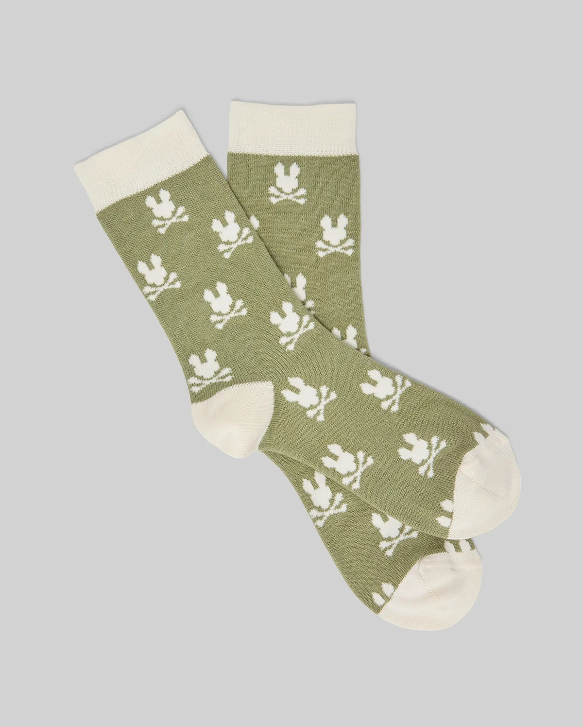 The Psycho Bunny KIDS 2 PACK ALL OVER BUNNY DRESS SOCK - KO2200003 features green socks with white cuffs, heels, and toes, decorated with a repeating white skull and crossbones bunny-ear pattern on light gray, made from soft stretch Pima cotton blend.