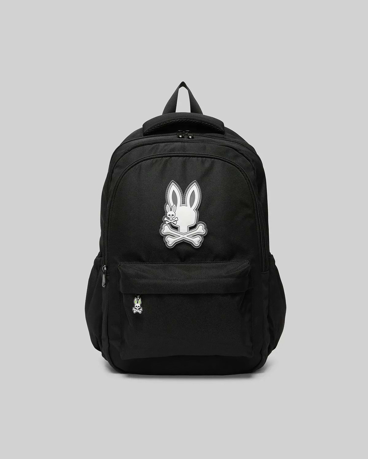 The Psycho Bunny KIDS PSYCHO BUNNY BACKPACK - KO2400004 displays a white cartoon bunny and crossbones emblem on the front pocket. Made from durable canvas with padded, adjustable straps, it is shown on a plain gray background.