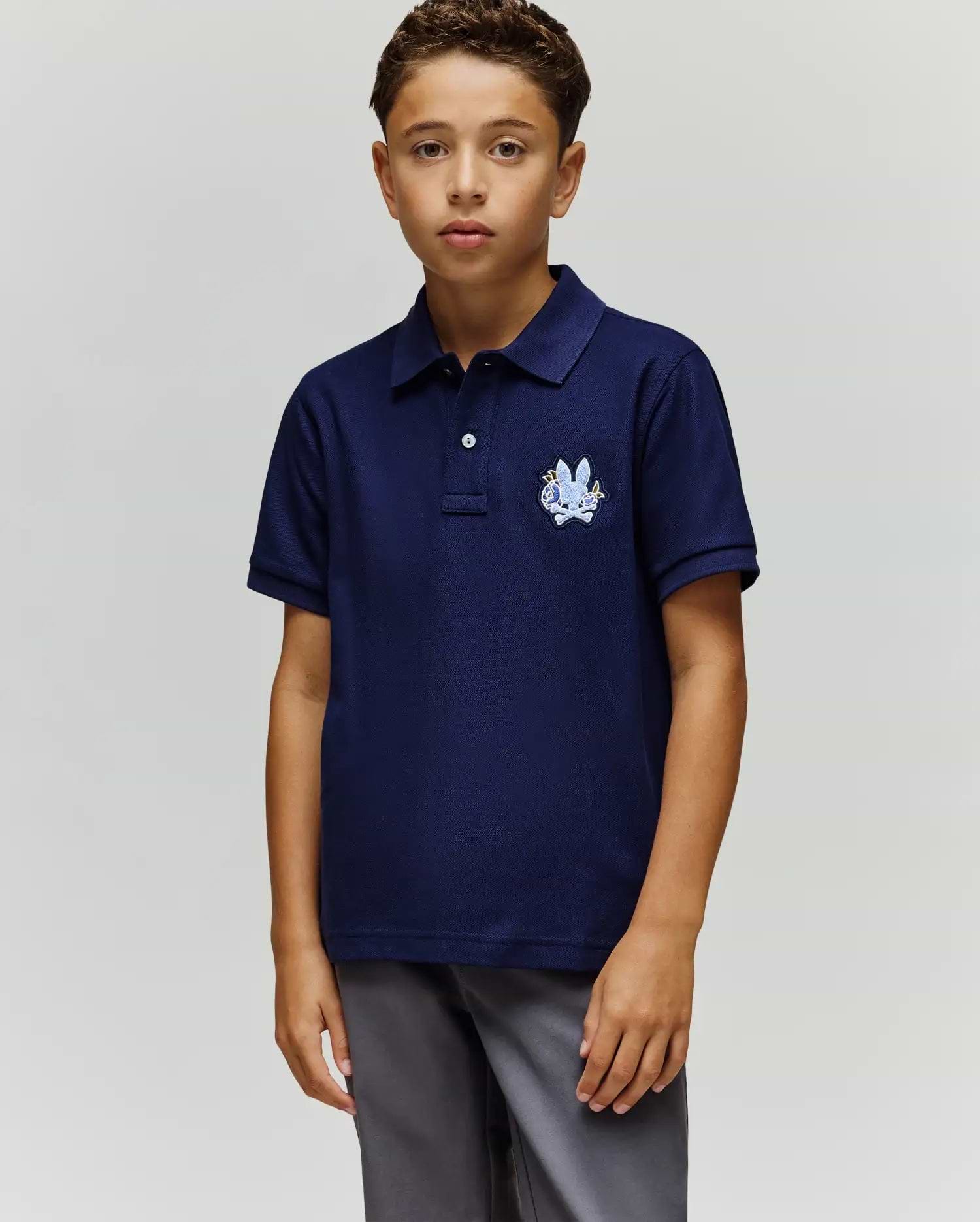 A boy with short curly hair and a neutral expression stands against a plain background, wearing the Psycho Bunny KIDS AUGUSTE PIQUE POLO - KT0100033 in navy blue with a crest on the left chest and gray pants, his left arm relaxed by his side. (1)