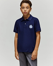 A boy with short curly hair and a neutral expression stands against a plain background, wearing the Psycho Bunny KIDS AUGUSTE PIQUE POLO - KT0100033 in navy blue with a crest on the left chest and gray pants, his left arm relaxed by his side. (1)