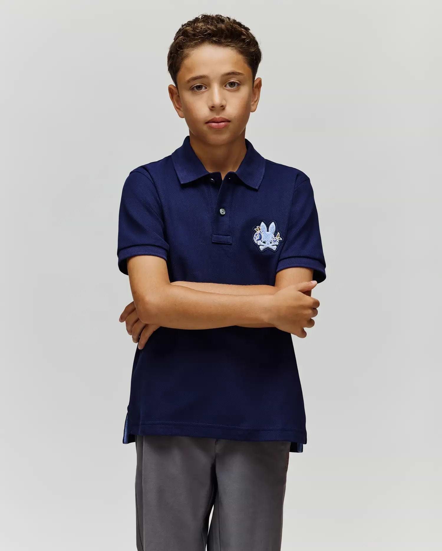 A boy with short curly hair stands against a plain background, arms crossed and neutral-faced, wearing the Psycho Bunny KIDS AUGUSTE PIQUE POLO - KT0100033 in varsity-style navy blue and gray pants. (3)