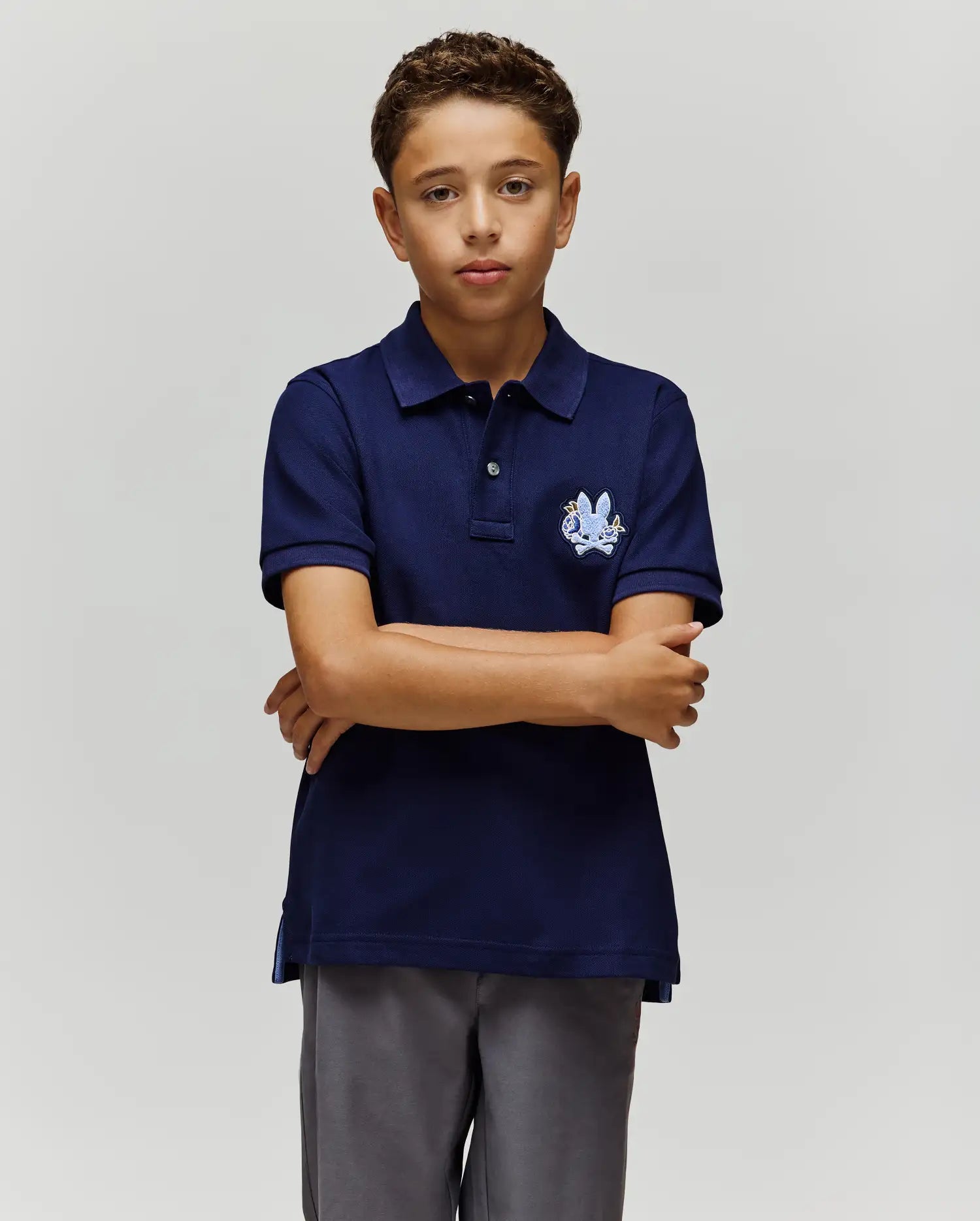 A boy with short curly hair stands against a plain background, arms crossed and neutral-faced, wearing the Psycho Bunny KIDS AUGUSTE PIQUE POLO - KT0100033 in varsity-style navy blue and gray pants. (3)