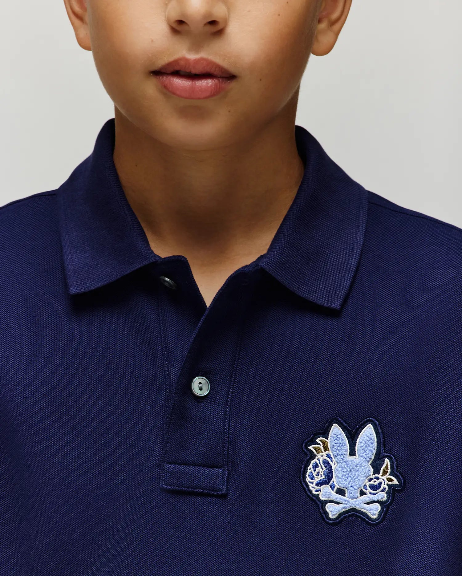 A close-up shows a child wearing the Psycho Bunny KIDS AUGUSTE PIQUE POLO (KT0100033) in navy blue with a light blue bunny and crossbones on the left chest. Only the lower half of the child's face is visible. (2)