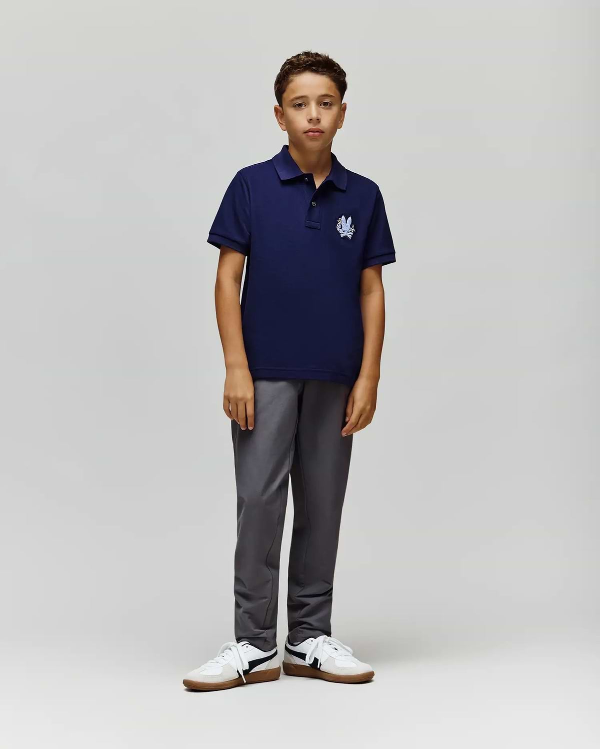 A young boy with short curly hair and a neutral expression wears the Psycho Bunny KIDS AUGUSTE PIQUE POLO - KT0100033 in navy Pima cotton with a white logo, paired with gray pants and white sneakers against a plain background. (4)