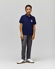 A young boy with short curly hair and a neutral expression wears the Psycho Bunny KIDS AUGUSTE PIQUE POLO - KT0100033 in navy Pima cotton with a white logo, paired with gray pants and white sneakers against a plain background. (4)