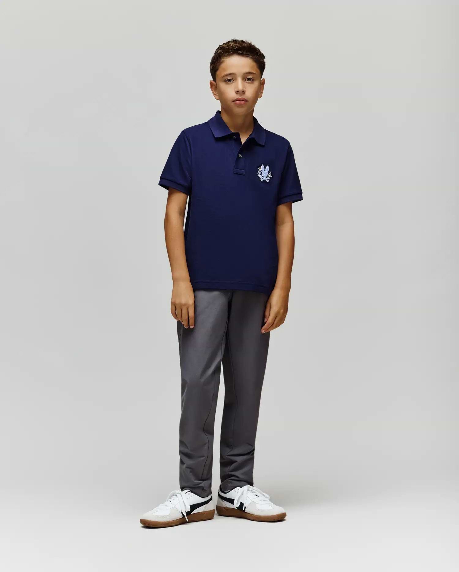 A young boy with short curly hair and a neutral expression wears the Psycho Bunny KIDS AUGUSTE PIQUE POLO - KT0100033 in navy Pima cotton with a white logo, paired with gray pants and white sneakers against a plain background. (4)