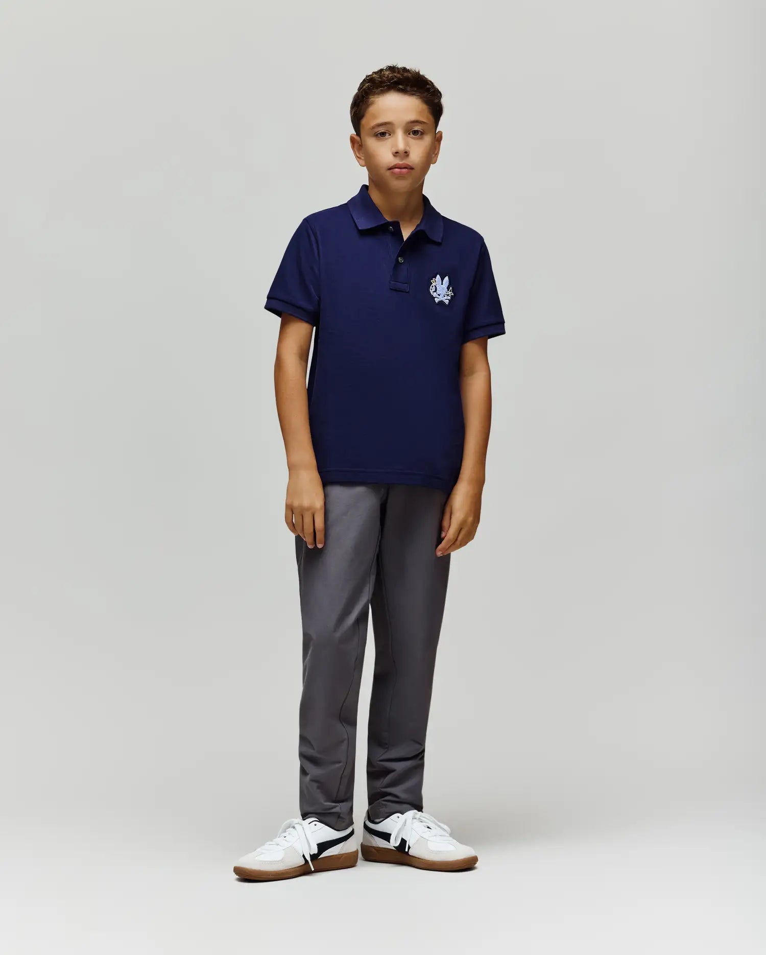 A young boy with short curly hair and a neutral expression wears the Psycho Bunny KIDS AUGUSTE PIQUE POLO - KT0100033 in navy Pima cotton with a white logo, paired with gray pants and white sneakers against a plain background. (4)