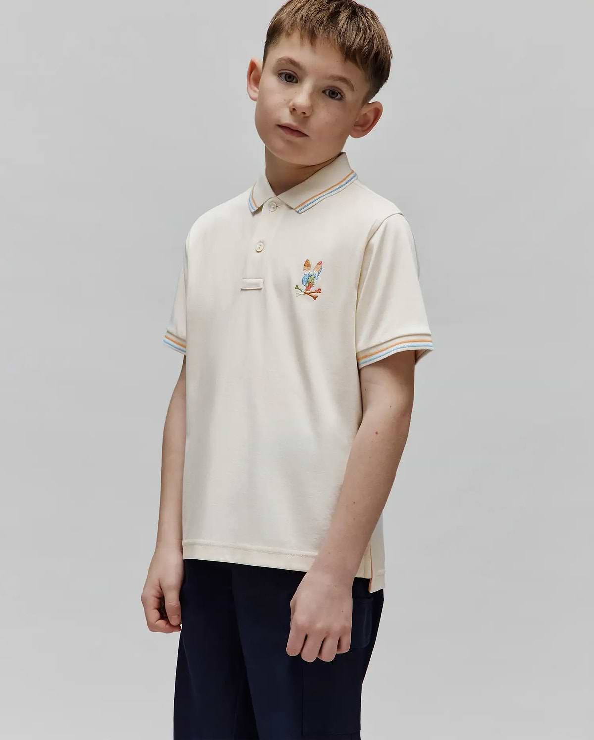 Kids Ara Pique Polo in cream pique knit, featuring a small embroidered Psycho Bunny skull with golf clubs on the left chest, and striped collar and sleeve cuffs in orange, light blue, and white.