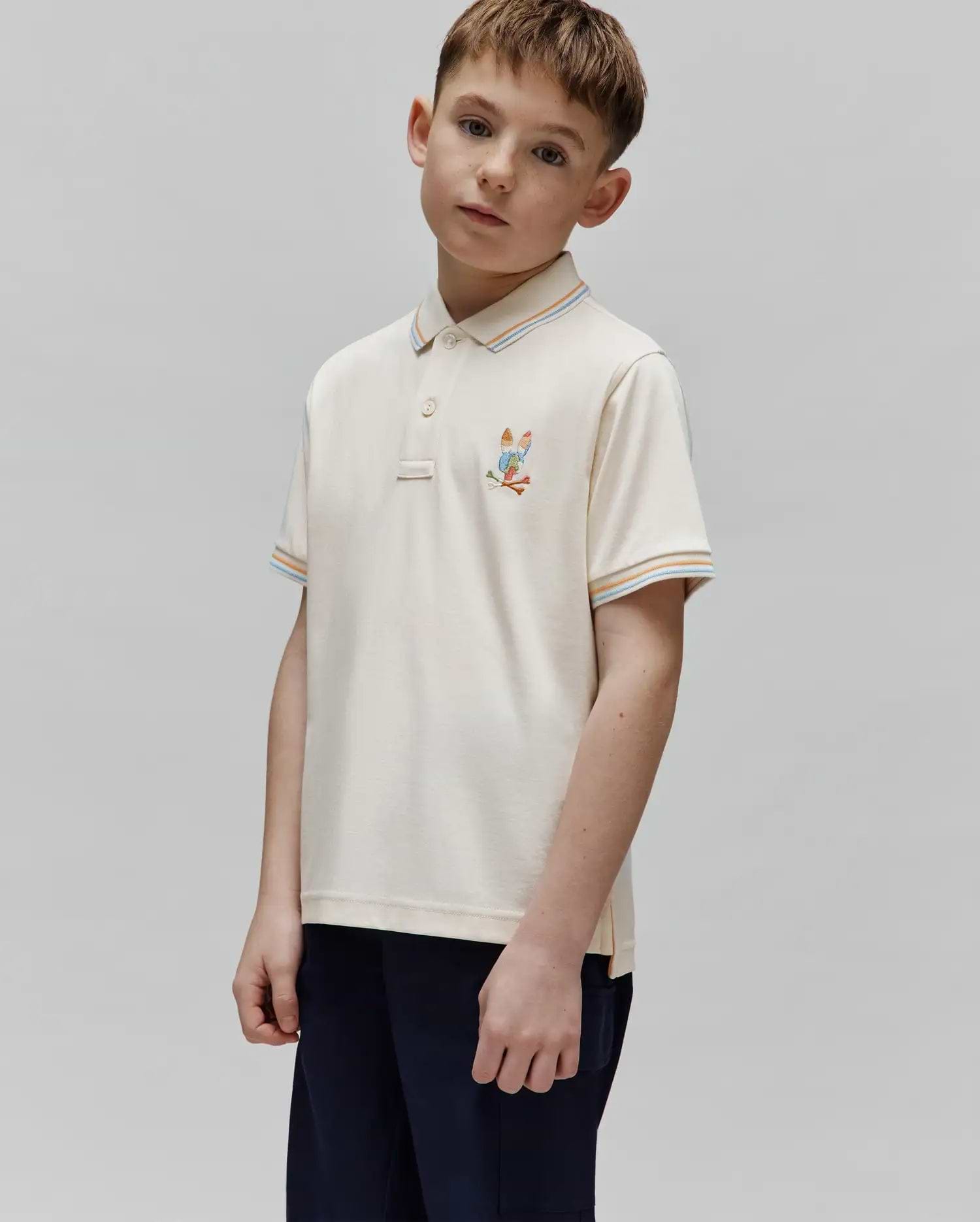 Kids Ara Pique Polo in cream pique knit, featuring a small embroidered Psycho Bunny skull with golf clubs on the left chest, and striped collar and sleeve cuffs in orange, light blue, and white.