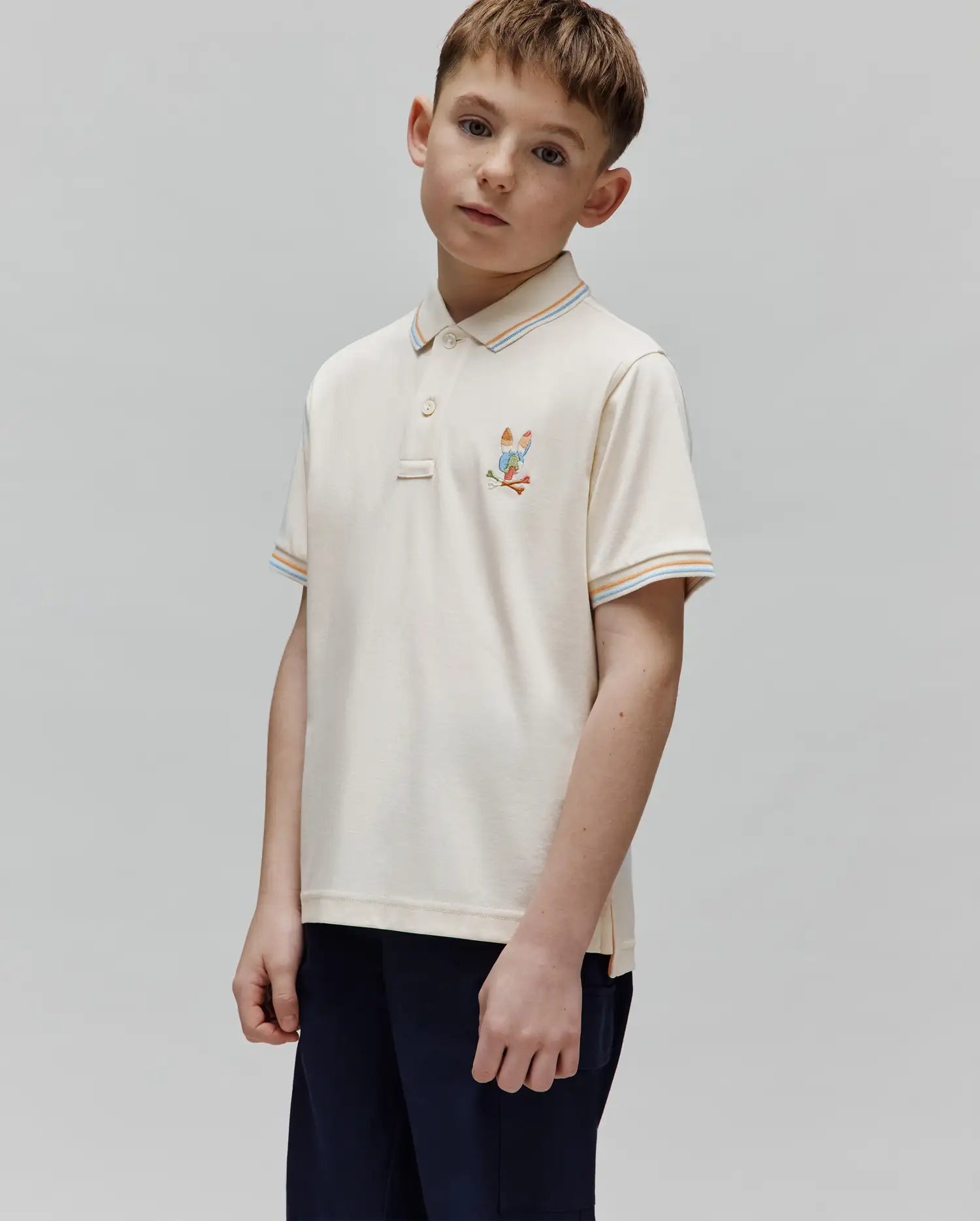 Kids Ara Pique Polo in cream pique knit, featuring a small embroidered Psycho Bunny skull with golf clubs on the left chest, and striped collar and sleeve cuffs in orange, light blue, and white.