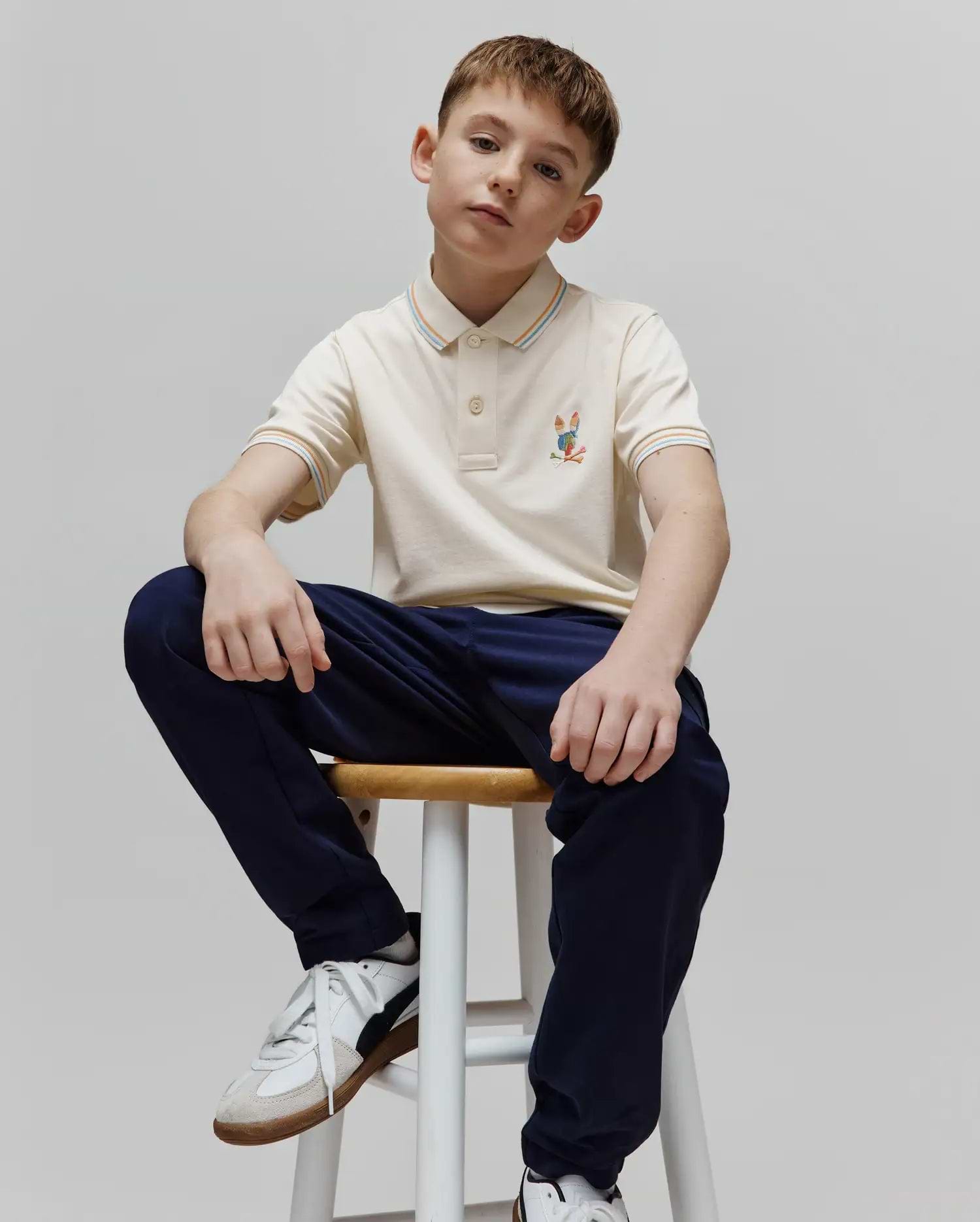 Kids Ara Pique Polo in cream pique knit, classic fit, with a colorful embroidered Psycho Bunny skull logo on the left chest and striped collar and cuffs featuring orange, blue, and white accents.