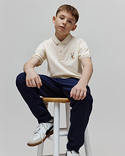 Kids Ara Pique Polo in cream pique knit, classic fit, with a colorful embroidered Psycho Bunny skull logo on the left chest and striped collar and cuffs featuring orange, blue, and white accents.