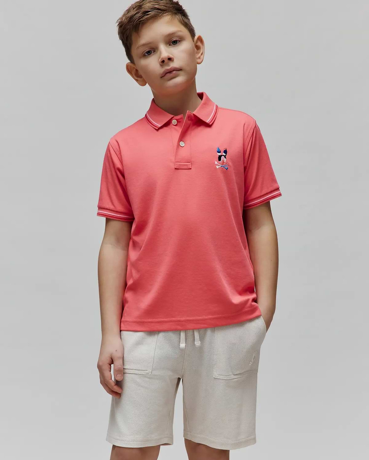 Kids Ara Pique Polo in coral, classic fit, pique fabric. Features a small, multi-color embroidered Psycho Bunny skull logo on the left chest and white tipping on the ribbed collar and cuffs.