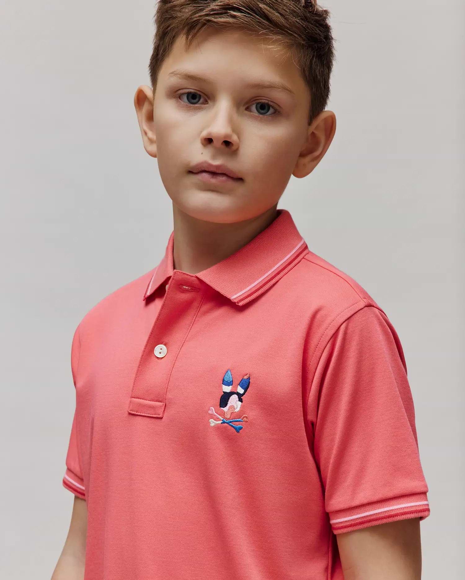 Kids Ara Pique Polo in coral pink, featuring a classic fit, pique knit fabric, ribbed collar and cuffs with white and light pink double stripes, and an embroidered multi-color Psycho Bunny skull logo on the left chest.